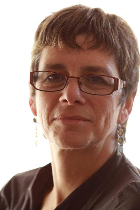 A white woman with short hair, wearing glasses, long earrings and a brown top
