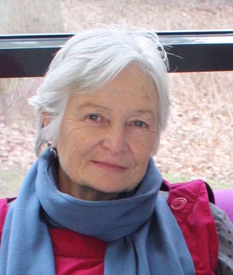 A white woman with short grey hair wearing a red coat and blue scarf