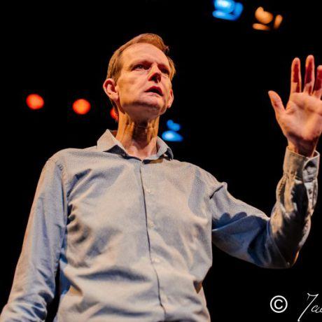 a white man with short brown hair, wearing a blue shirt and gesturing with his hand