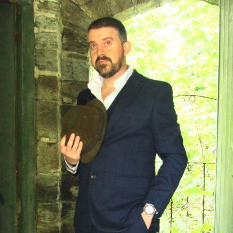 a white man with short dark hair and beard, wearing a suit and holding a hat