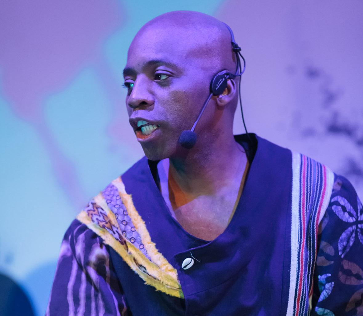 a black man wearing a black and gold tunic and playing a drum