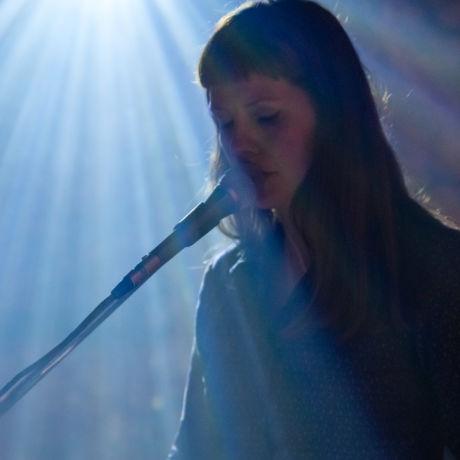 a white woman with long dark hair singing into a microphone on stage. there is a strong shaft of light.