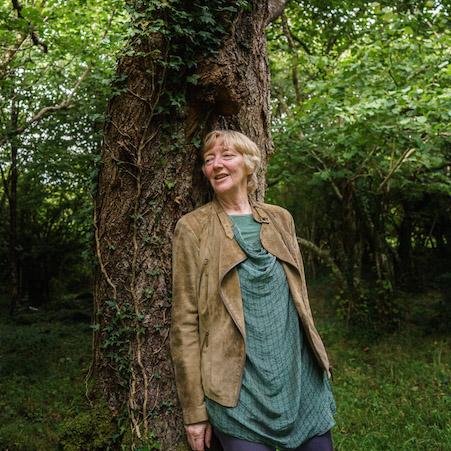 a white woman with short blond hair, wearing a green tunic and light brown jacket. leaning against a tree in the woods
