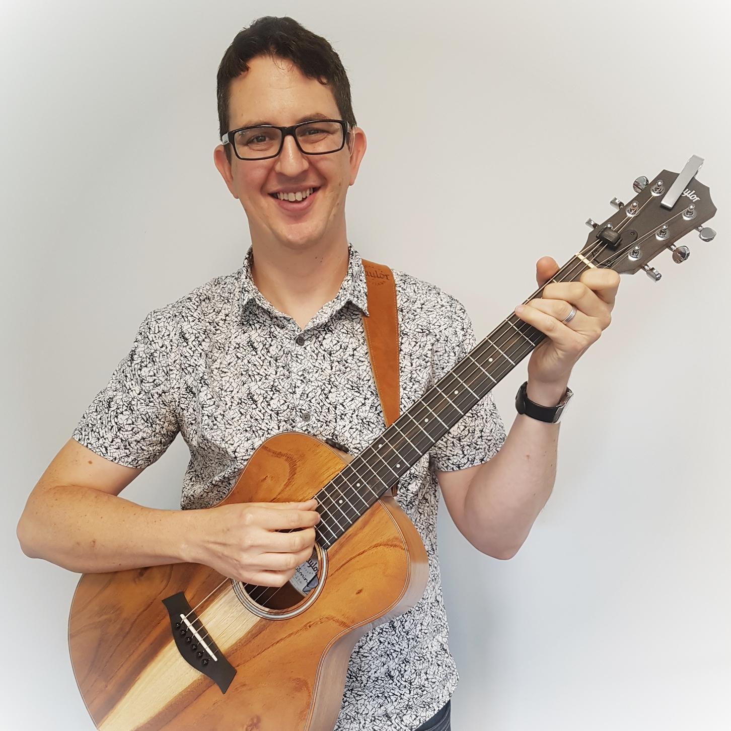 A white man with short dark hair, wearing glasses and a white and black shirt and holding a guitar