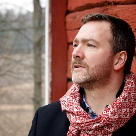 a white man with short brown hair and a beard, wearing a red and white scarf