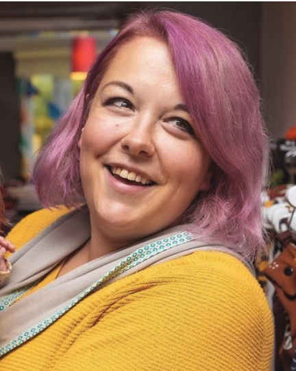 a white woman with shoulder length pink hair wearing a yellow jumper