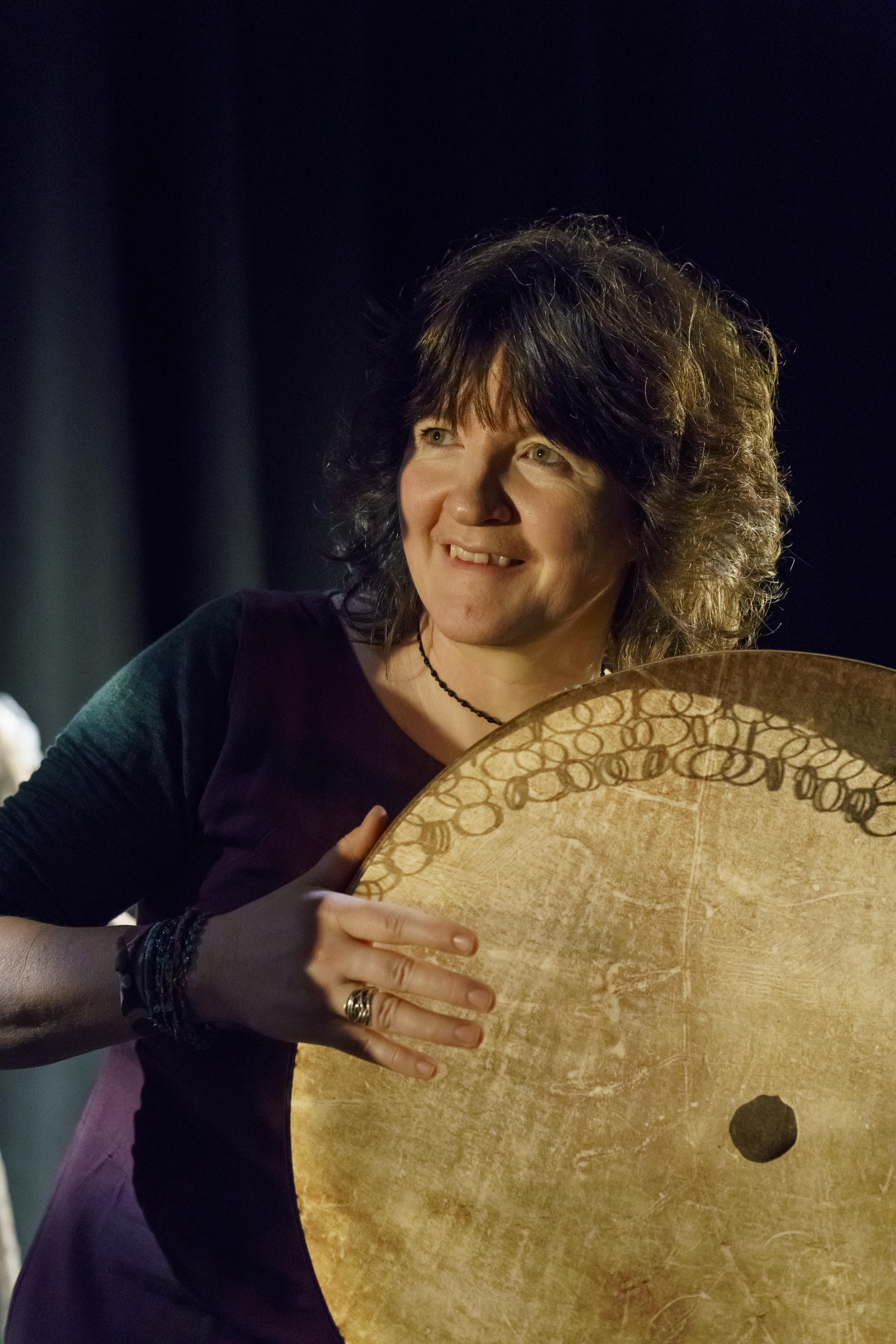 a white woman with shoulder length brown hair, playing a large drum