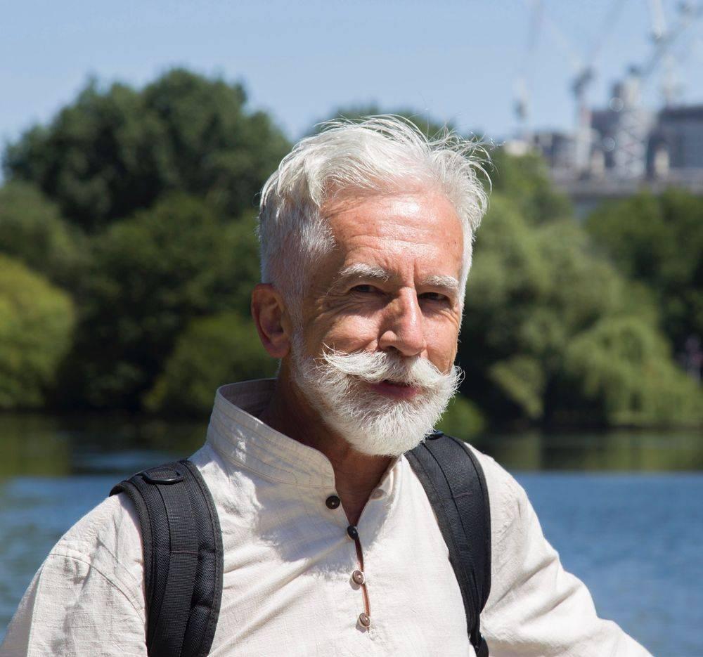 A white man with white hair and beard in front of a lake