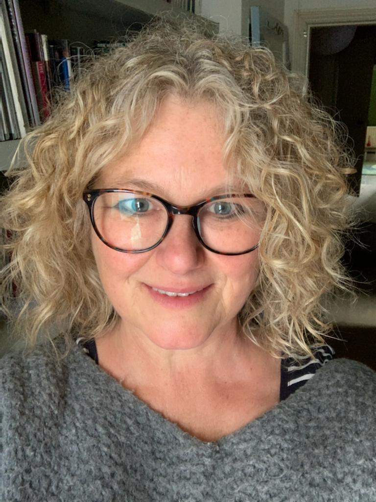 a white woman with shoulder length curly blond hair and wearing dark rimmed glasses