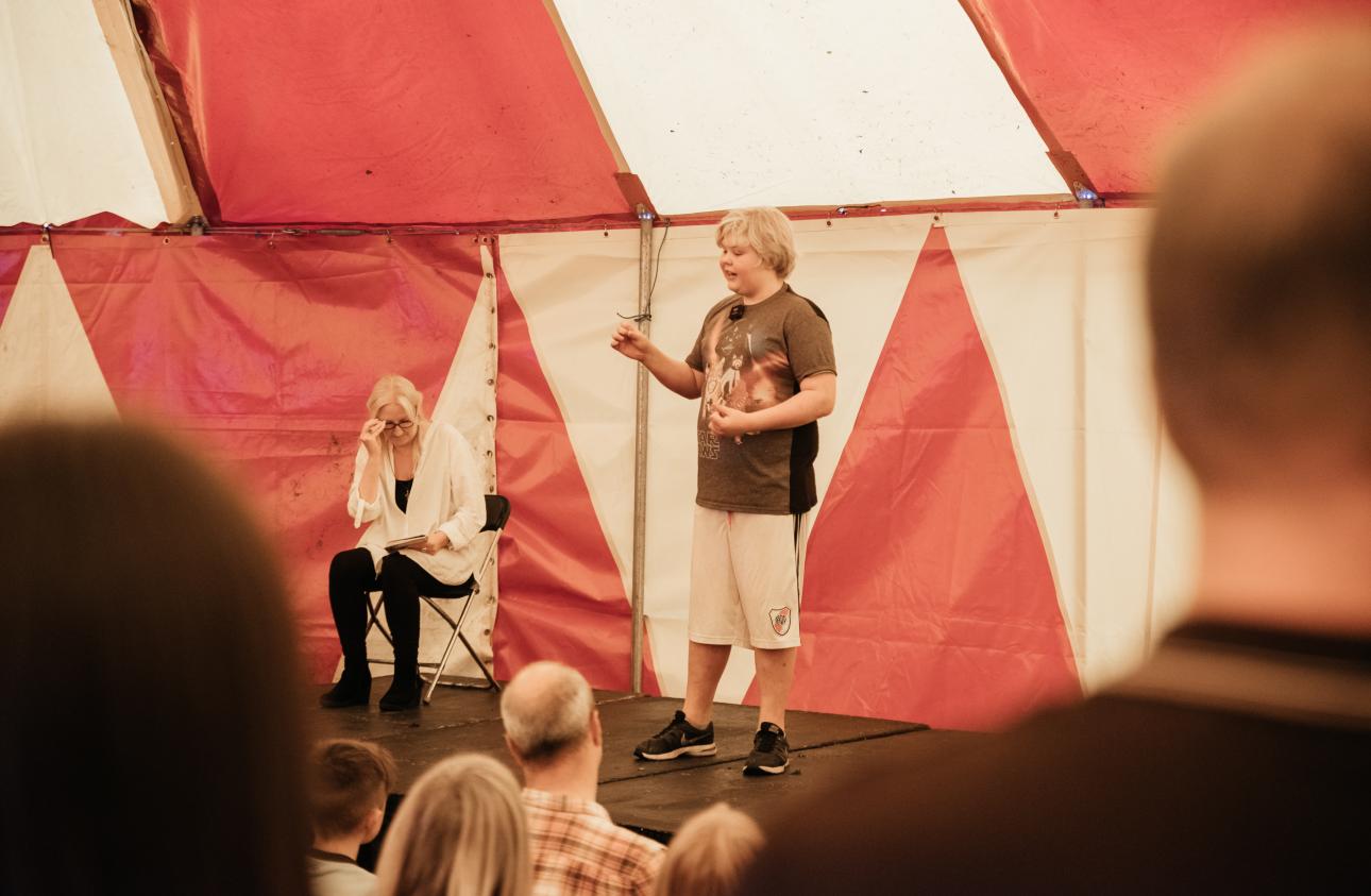 a young person tell their story on stage in a red and yellow big top tent