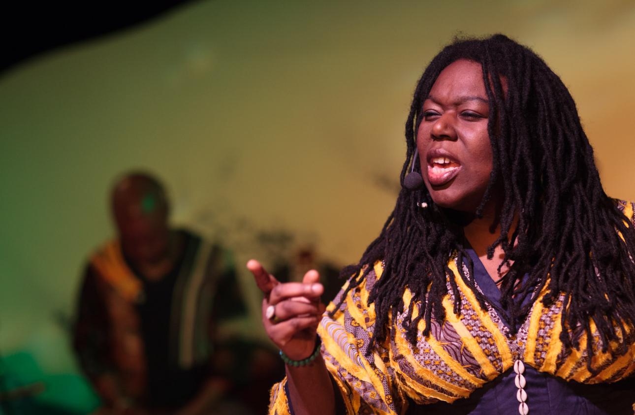 Jan Blake seated and performing, she is pointing forwards