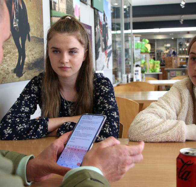 a man holding a phone sits away from us facing 2 young girls in a cafe