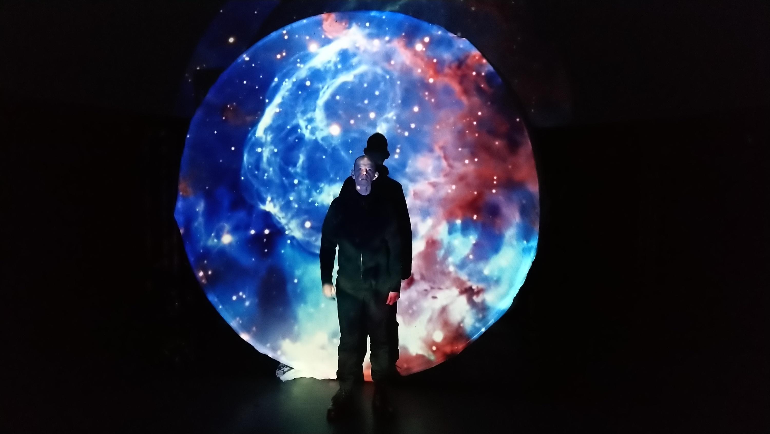 Storyteller Daniel Morden wearing dark clothes in a dark space stands in front of a disc that has images of the night sky projected onto it