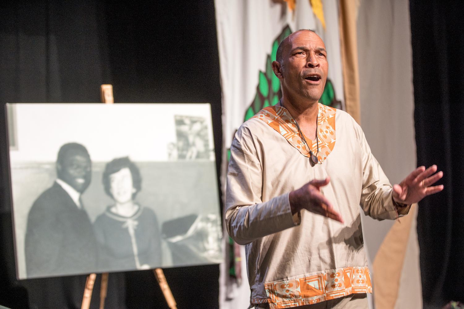 Phil Okwedy is standing on a stage looking out to the audience, with his arms raised up in front of him, and is stood in front a photograph of his parents. This is the only photo he has of his Nigerian Father and his Welsh mother before his father moved back to Nigeria.