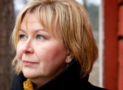 A white woman with short blond hair wearing a dark coat and yellow scarf