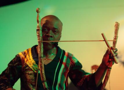 a black man wearing a black and gold tunic and playing the dodo mouth bow