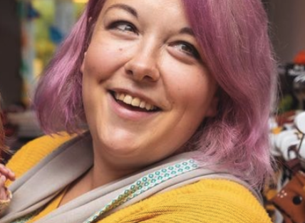 a white woman with shoulder length pink hair wearing a yellow jumper