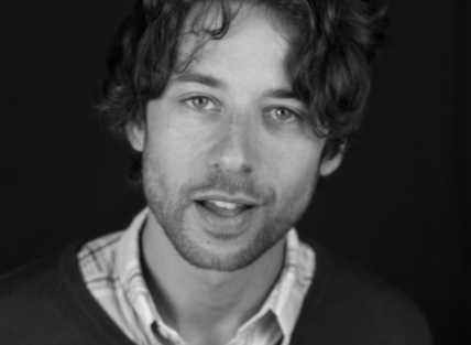 a young white man with curly hair and a beard. wearing a shirt and v neck jumper