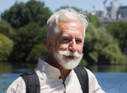 A white man with white hair and beard in front of a lake