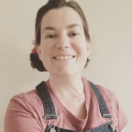 a white woman with dark hair, wearing a pink top and dungarees