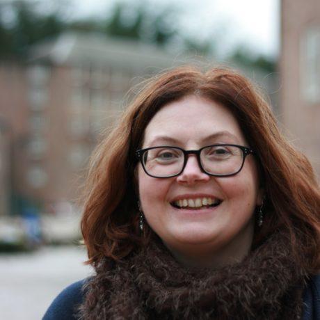 A woman with shoulder length auburn hair, wearing glasses, blue jumper and brown scarf