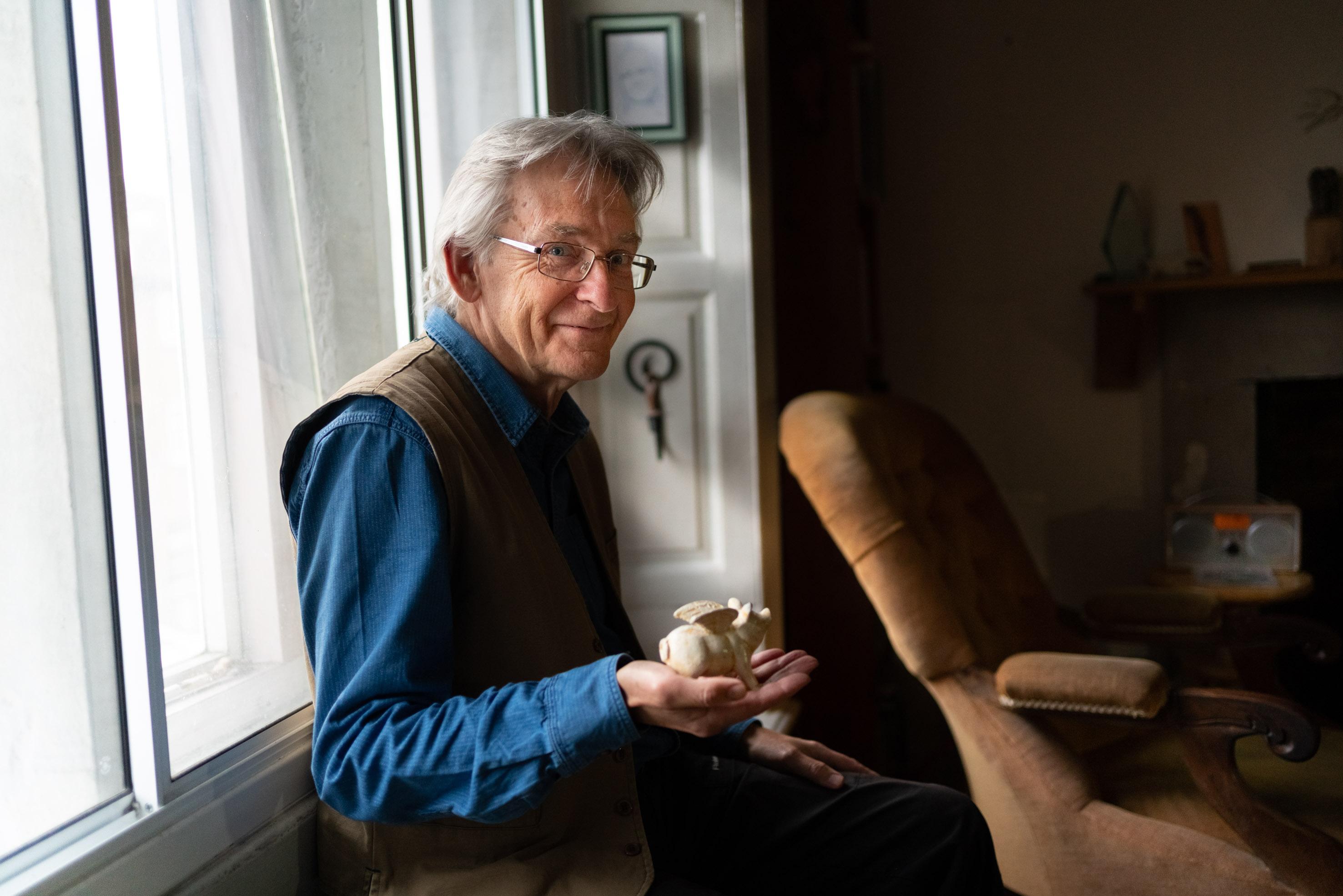 A white man with grey hair, wearing a blue shirt and waistcoat holding a ceramic winged pig