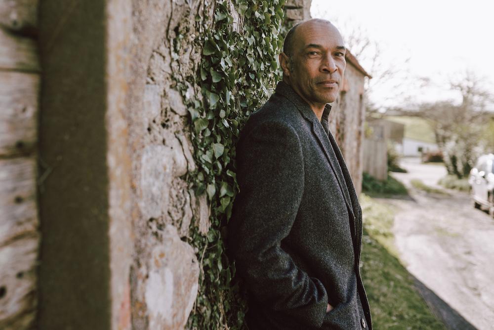 a black man with cropped hair, wearing a winter coat and leaning against a wall