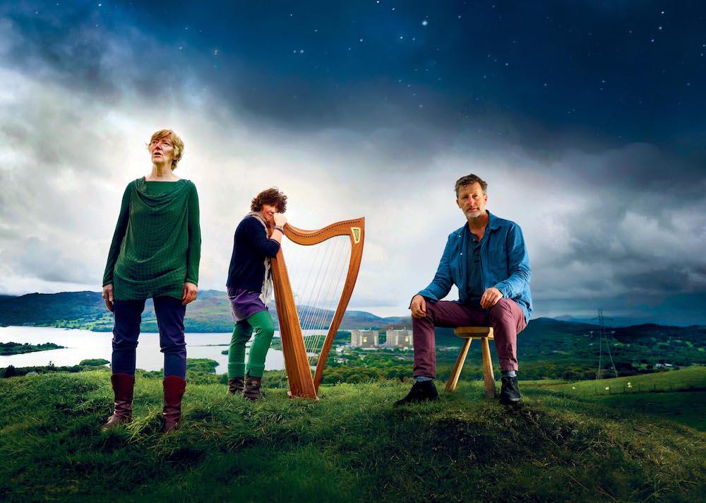 3 artists stand on top of a hillm one sitting on a stool, one holding a harp, a dramatic sky