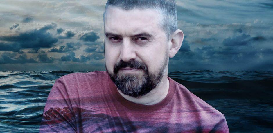 a head and shoulders photo of Gwilym Morus-Baird, a white man with short hair and beard wearing a pink t-shirt. Against a background of a stormy sea.