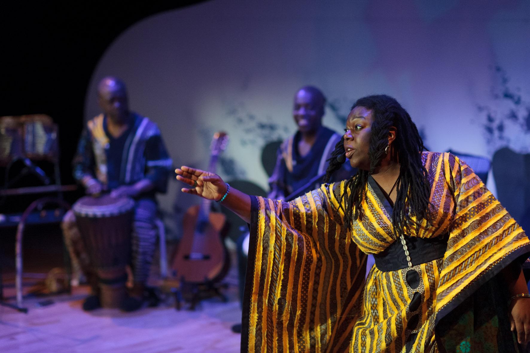 storyteller Jan Blake wearing a gold and yellow costume stretches out her hand, musicians Raymond & Kouame Sereba can be seen in the background
