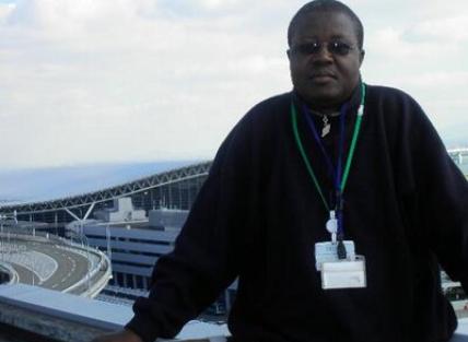 photograph of Calistus Wachana standing in front of a large building perhaps a stadium or airport. A young black man with short hair and wearing glasses