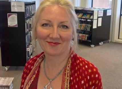 A white woman with blond hair tied back, wearing a red jacket and necklace