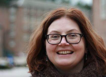 A woman with shoulder length auburn hair, wearing glasses, blue jumper and brown scarf