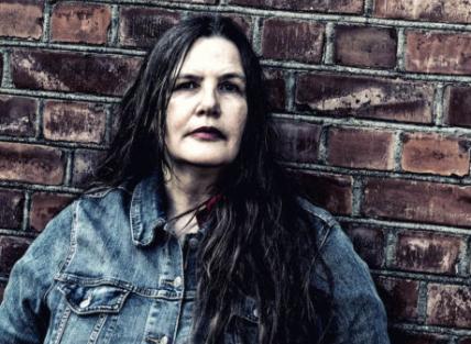 A white woman with long dark hair wearing a denim jacket and leaning against a brick wall