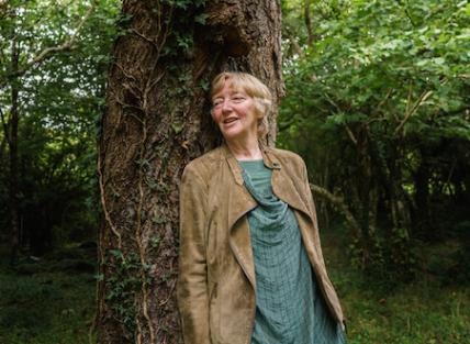 a white woman with short blond hair, wearing a green tunic and light brown jacket. leaning against a tree in the woods