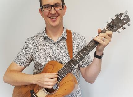 A white man with short dark hair, wearing glasses and a white and black shirt and holding a guitar